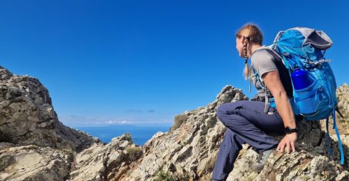 La dott.ssa Ferrari seduta su rocce di montagna durante una pausa mindful, con zaino blu e panorama aperto sotto un cielo limpido.