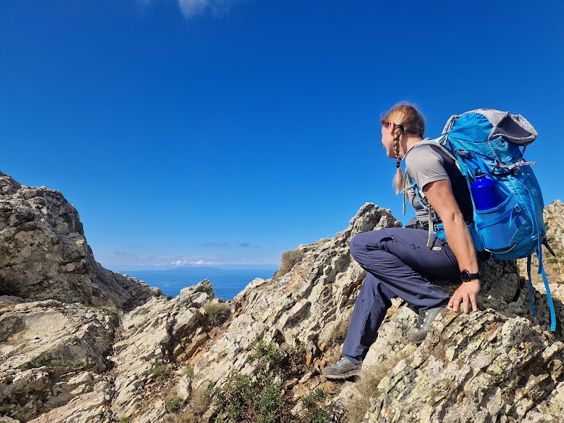 La dott.ssa Ferrari seduta su rocce di montagna durante una pausa mindful, con zaino blu e panorama aperto sotto un cielo limpido.